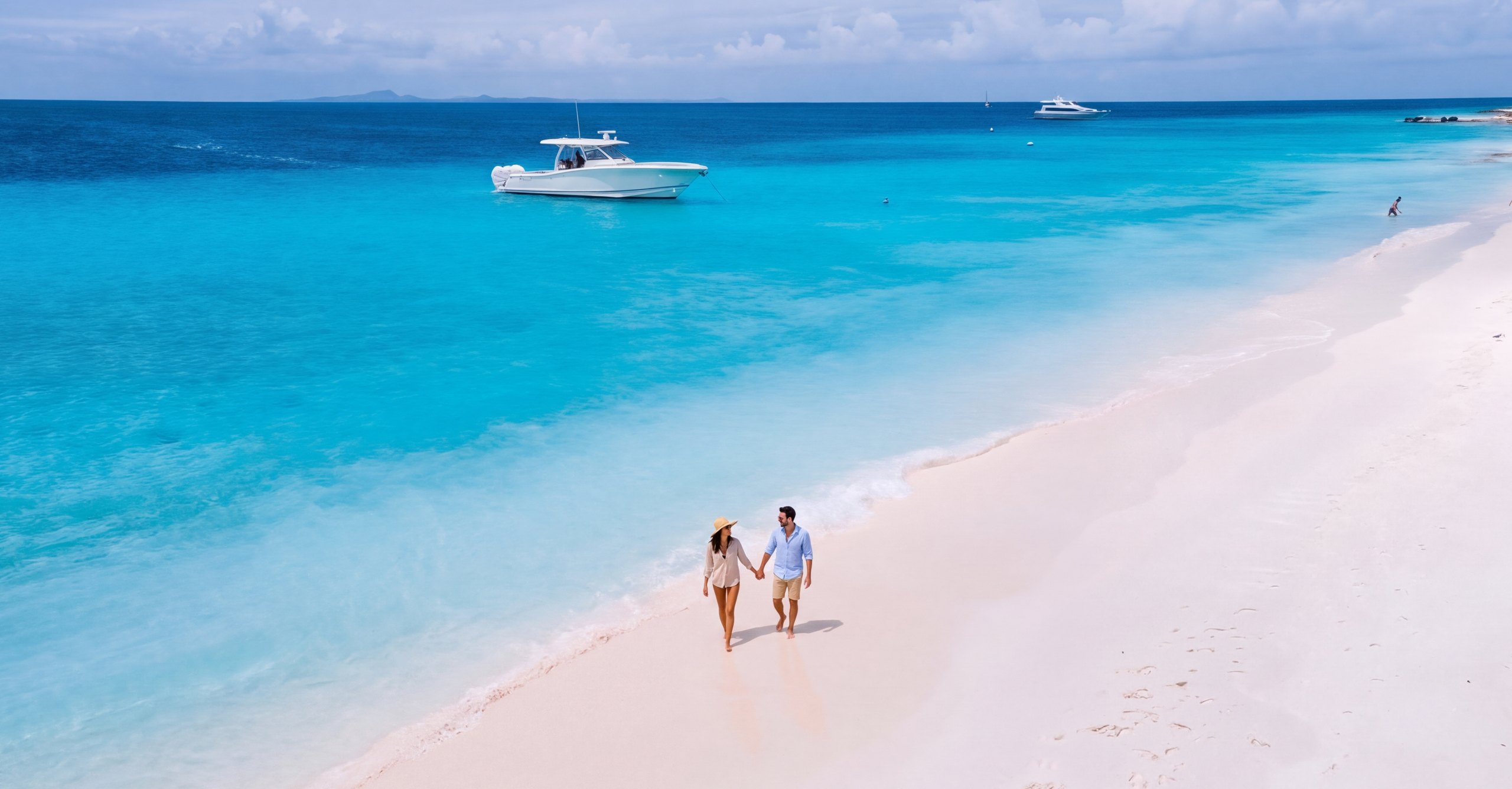 A couple walking hand in hand along a white sand beach, with a private boat anchored in turquoise water behind them