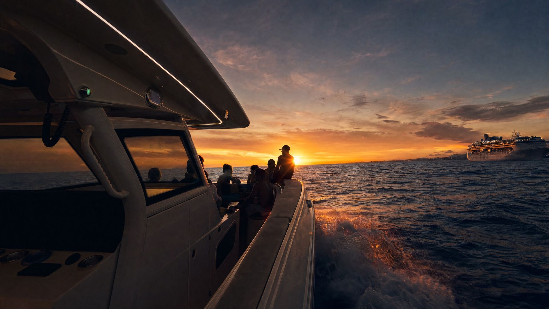 Guests on the bow of a private boat cruising toward the sunset off Curaçao, with a cruise ship on the horizon